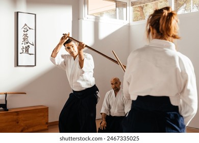 Two martial arts instructors, dressed in white gi with black hakama, demonstrate defensive jo staff maneuvers under the observant eye of a seated student. Text: "Aikido" - Powered by Shutterstock