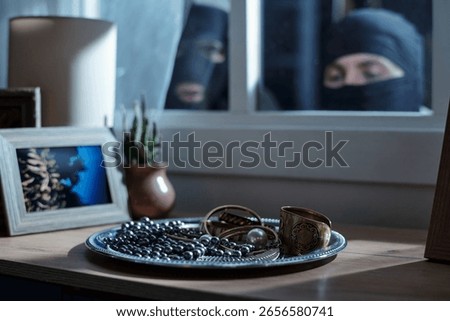 Two masked men peering through window observing jewelry and valuables on wooden table, framed photograph and potted cactus visible in foreground, suggesting burglary in progress