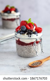 Two mason jars with chia pudding and fresh berry fruits and mint on a white background with a wooden spoon with chia seeds in front