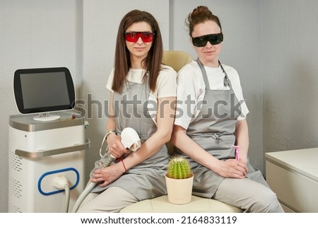 two masters of depilation sit on a couch in goggles. one holding a laser handpiece, the other a pink razor
