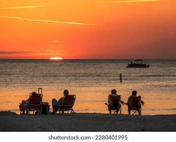 Two mature couples in beach chairs watch the sun flatten out moments before it disappears over the Gulf of Mexico, while passengers on a small tour boat take in the view offshore. Southwest Florida.