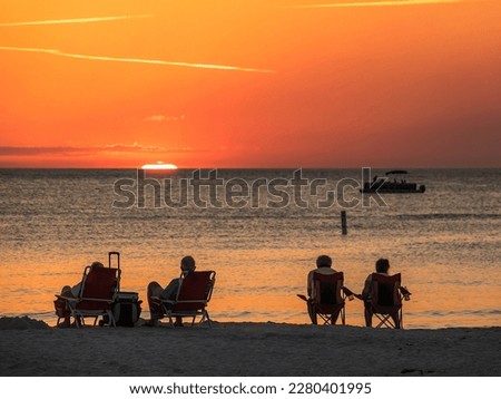 Two mature couples in beach chairs watch the sun flatten out moments before it disappears over the Gulf of Mexico, while passengers on a small tour boat take in the view offshore. Southwest Florida.