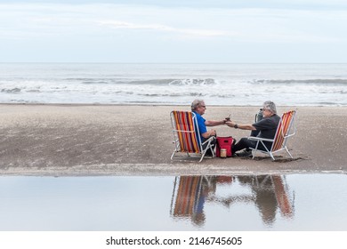 Two Mature Man With Gray Hair Sitting On A Beach Chair Drinking Mate All Reflected In The Water. Behind The Sea Waves.