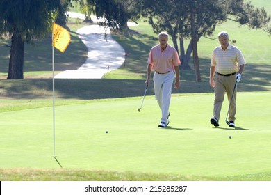 Two Mature Men Playing Golf, Approaching Balls Near Flag On Putting Green, Smiling