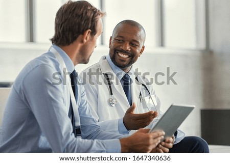 Two mature smiling doctors having discussion about patient diagnosis, holding digital tablet. Representative pharmaceutical discussing case after positive result with happy doctor about new medicine.