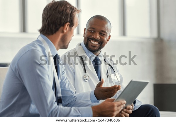 Two mature smiling doctors having discussion
about patient diagnosis, holding digital tablet. Representative
pharmaceutical discussing case after positive result with happy
doctor about new medicine.