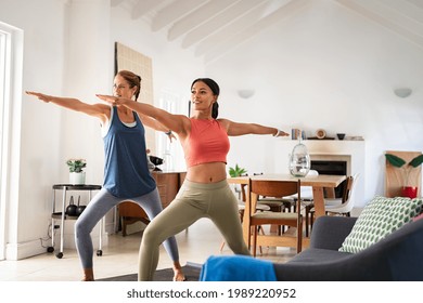 Two mature women doing warrior pose at home with stretched arms. Healthy mature woman doing yoga exercise with mixed race friend in living room. Middle aged lady in sportswear exercising at home. - Powered by Shutterstock