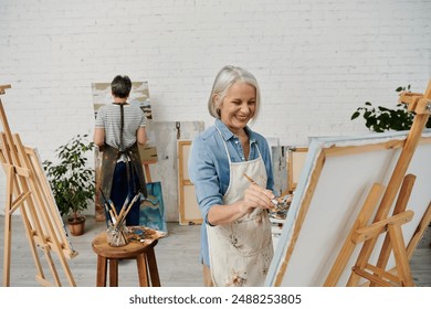 Two mature women are painting in an art studio. One woman is smiling as she holds a paintbrush and palette. - Powered by Shutterstock