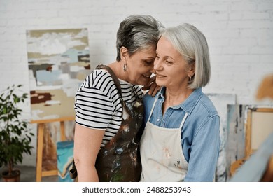 Two mature women in paint-splattered aprons share a tender moment in an art studio, faces full of love and joy - Powered by Shutterstock