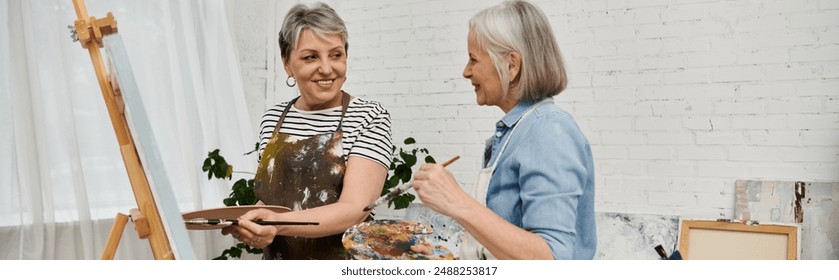 Two mature women stand in an art studio, one holding a paintbrush and palette, while the other smiles and holds a paintbrush. - Powered by Shutterstock