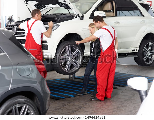 two mechanical employees of the car service workshop\
make some repairs at the blonde female customer car and one of them\
show her something at the tires or rims whats to repair on the\
jacked car