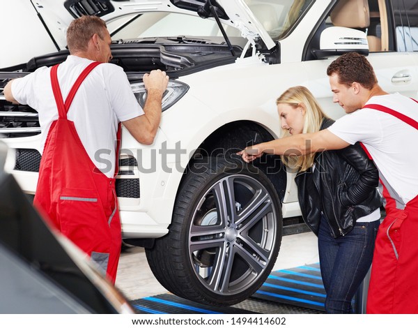 two mechanical employees of the car service workshop\
make some repairs at the blonde female customer car and one of them\
show her something at the tires or rims whats to repair on the\
jacked car