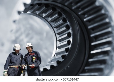 Two mechanics, workers in front of giant gears axle, as with steel industrial works
