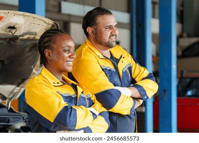 Two mechanics in yellow and blue uniforms, smiling confidently, arms crossed, standing in garage, with car and open hood in background, teamwork in automotive repair. Service work in car repair shop - Powered by Shutterstock