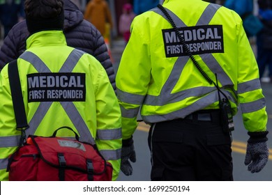 Two Medical First Responders Walking In A Street Among A Crowd Of People.One Of The EMTs Is Carrying A Red First Aid Bag.The Emergency Personnel Have Bright Yellow Reflective Jackets With Grey Stripes