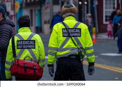 Two Medical First Responders Walking In A Street With People In The Background. The Officers Are Wearing A Bright Yellow Reflective Coat With Grey Stripes And A Red First Aid Bag. 