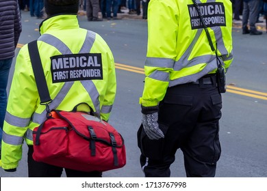 Two Medical Responders Walking In A Street With Bright Yellow Coats With The Words Medical First Responder On A Black Background With Grey Lettering. One Medical Officer Is Carrying A First Aid Bag.
