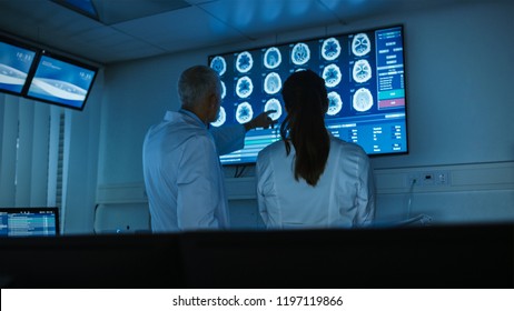 Two Medical Scientists / Neurologists, Talking and Pointing at Wall TV Showing CT / MRI Brain Scans in Modern Laboratory. Research Scientists Making New Discoveries in the fields of Neurophysiology