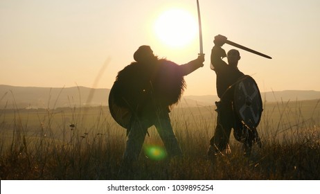 Two medieval warriors Viking are fighting with swords and shields in the meadow. Medieval Reenactment. Contre-jour