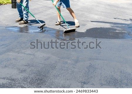 Two men applying sealer on asphalt driveway with brush and squeegee on sunny day