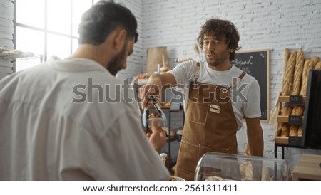 Two men in a bakery with one serving as a baker and the other as a customer, exchanging baked goods in an indoor shop setting.