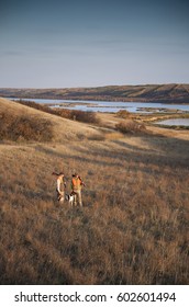 Two Men, Bird Hunters, With Shotguns, Carrying The Day's Bag Of Dead Birds, And A Spaniel Dog