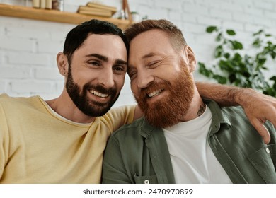 Two men, both with beards, relax on a couch and hugging at home