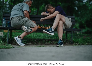 Two men in casual attire engage in a strategic chess game on a park bench, surrounded by greenery, depicting a relaxed and thoughtful atmosphere. - Powered by Shutterstock