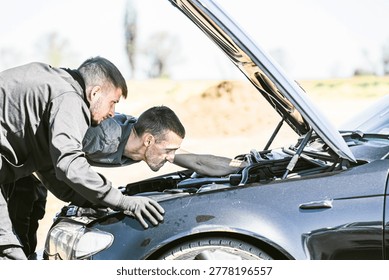 Two men check the engine components of a car with the hood raised in an outdoor environment, surrounded by trees and dirt in the background