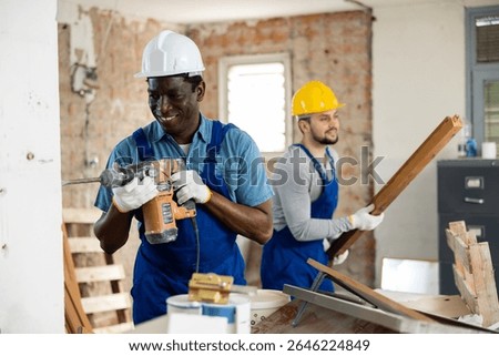 Two men construction workers drilling wall with pneumatic hammer drill and carrying wooden planks on indoor building construction site