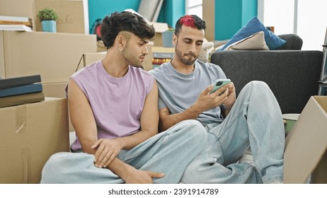 Two men couple using smartphone sitting on floor at new home