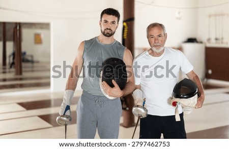 Two men of different ages after sparring at a fencing club. Elderly man and a young man stand in the gym, they hold swords and safety helmets after fencing training