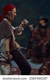 Two men, dressed like medieval prated raising glasses with rum, drinking again vintage green background in dimly-lit room. Concept of camaraderie, history, adventure, achievement
