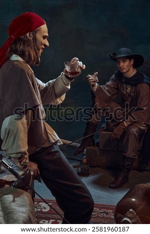 Two men, dressed like medieval prated raising glasses with rum, drinking again vintage green background in dimly-lit room. Concept of camaraderie, history, adventure, achievement