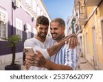 Two men embracing each other, smiling and taking a selfie with a smartphone while walking down a picturesque street, enjoying their love and time together