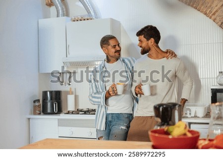 Two men embracing and smiling while drinking coffee in a modern kitchen, enjoying a quiet moment together
