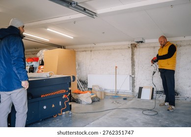 Two men are engaged in work inside a garage, with one using a power tool and the other observing near a table saw and assorted materials