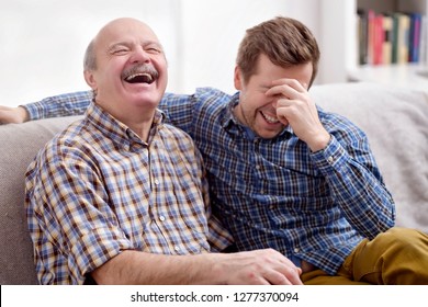 Two Men Father And Son Are Sitting On The Sofa In The Living Room At Home And Remember The Funny Events From The Past. They Laugh At The Joke. Good Family Relationships