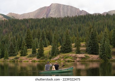 Two men fishing with a dog in a lake with a pine forest and mountains in the background - Powered by Shutterstock