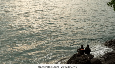 Two men fishing as a relaxing hobby, sitting on a large rock by the beach. Gentle waves and a peaceful ocean view create a serene and tranquil atmosphere for leisure. - Powered by Shutterstock