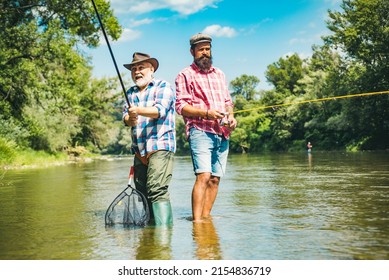 Two Men Friends Fishing. Flyfishing Angler Makes Cast, Standing In River Water. Old And Young Fisherman.