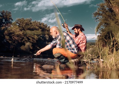 Two Men Friends Fishing. Flyfishing Angler Makes Cast, Standing In River Water. Old And Young Fisherman.