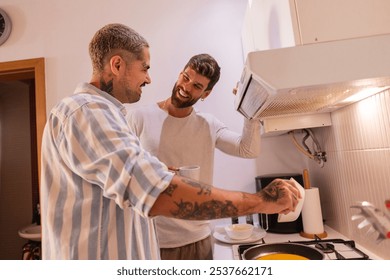 Two men, a gay couple, are cooking together in their kitchen, one is holding a cup of coffee while the other is checking the cooker hood while preparing breakfast - Powered by Shutterstock