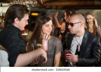 Two Men With The Girl In Chaindress Drink Cocktails At The Bar With Three Gils In The Background