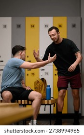 Two men are having a conversation in a locker room below the bright lockers. One man sits on a bench while the other stands with a water bottle. Their clothes suggest they just finished working out.