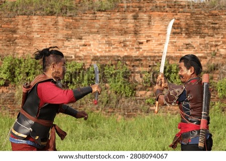 Two men in historical armor practice sword fighting outdoors against a weathered brick wall.