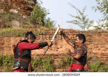 Two men in historical armor practice sword fighting outdoors against a weathered brick wall.