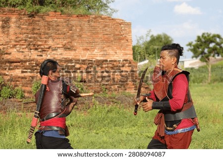 Two men in historical armor practice sword fighting outdoors by a weathered brick wall.