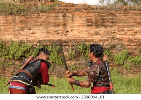 Two men in historical armor practice sword fighting outdoors against a weathered brick wall.