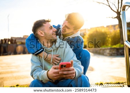 Two men hugging and laughing while using a smartphone outdoors, enjoying a sunny day together